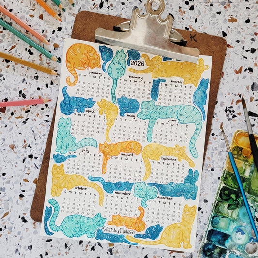 Calendar with cat illustrations on a clipboard with colorful pencils and art supplies.