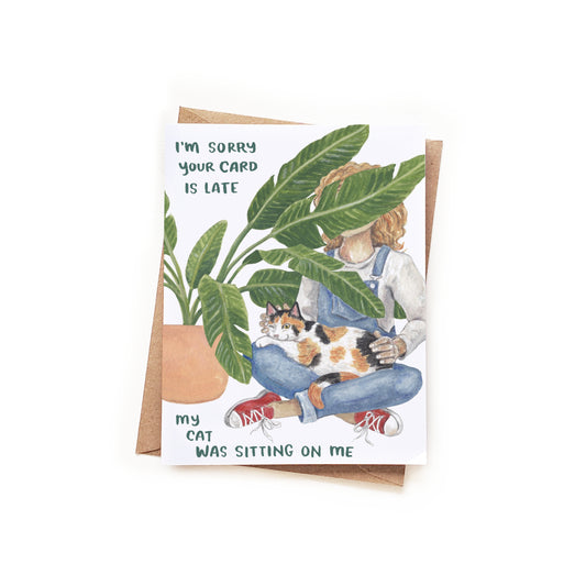 Card with illustration of a person holding a cat and text 'I'm sorry your card is late, my cat was sitting on me' on a white background.